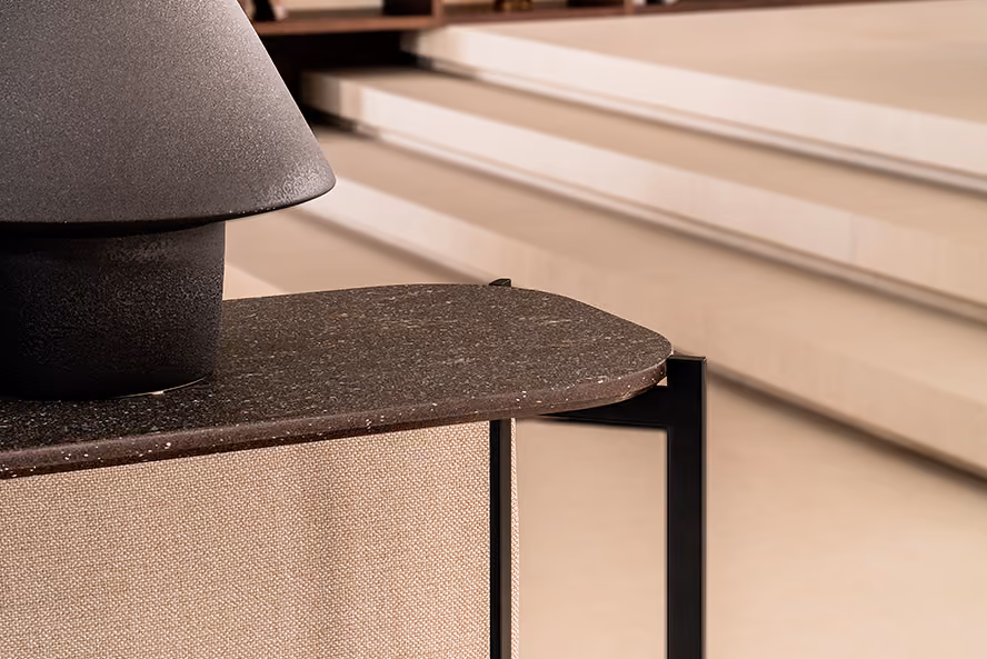 Close-up of a modern table corner with a dark textured stone top, supported by a black metal frame and beige fabric panel, with cream-colored steps in the background.