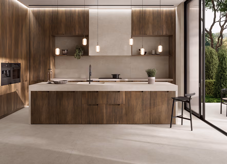 Modern kitchen with a large central island featuring a light beige countertop and wood-paneled cabinetry, pendant lights hanging above, a black faucet, a potted plant, and a black stool on the right side next to a large open glass door leading to a garden with trees and shrubs.
