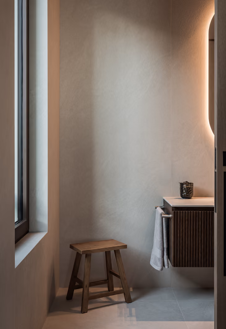 Minimalist bathroom corner with a wooden stool on a light tiled floor, a floating wooden vanity with vertical grooves, a white towel hanging from a bar, and a dark container on the vanity. Soft indirect light illuminates the space, and a window is on the left side.
