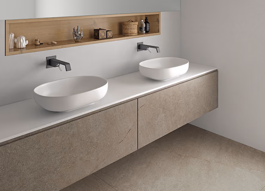 Modern bathroom with a floating vanity featuring two white oval vessel sinks and wall-mounted black faucets, a recessed wooden shelf with decorative items above the sinks, and beige stone tiles on the vanity and floor.