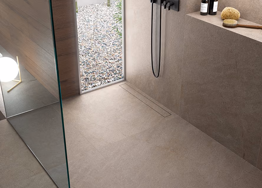 Modern minimalist shower area with beige stone tiles, a glass partition, and a black showerhead. A narrow window shows pebbles and plants outside. On a built-in shelf, there are two bottles, a round sponge, and a brush with a long handle.