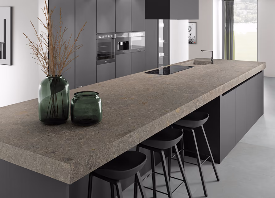 Modern kitchen with a large stone countertop island featuring an integrated sink and stovetop, accompanied by three black bar stools and two green glass vases with dried branches on the countertop, with tall grey cabinets and a window with sheer curtains in the background.