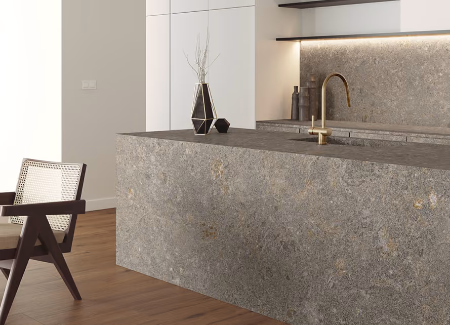 Modern kitchen with a large gray stone island countertop featuring a built-in sink and a gold faucet, a geometric vase with decorative sticks, and wooden floor, next to a wicker-back wooden chair.