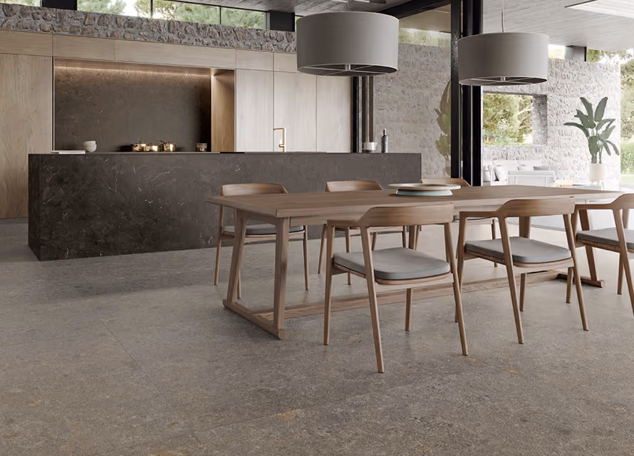 Modern dining area with a minimalist wooden table and six cushioned chairs, set on a stone-tiled floor with a dark stone kitchen island and wood cabinetry in the background, under large pendant lights and near large windows showing an outdoor stone wall and greenery.