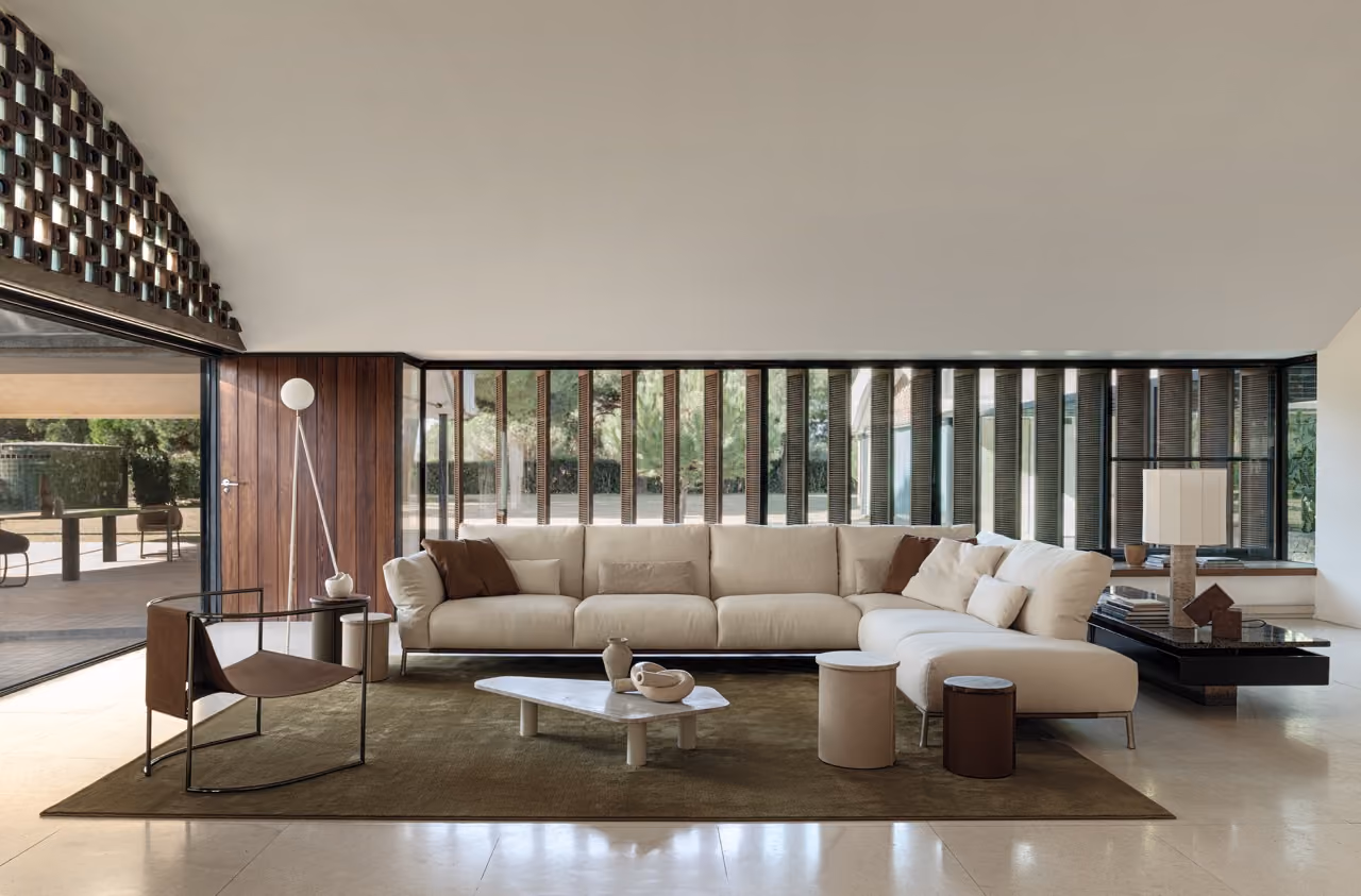 Modern living room with a large light beige sectional sofa, brown and beige cushions, two small round side tables, a black low rectangular coffee table with a lamp and decorative objects, a brown armchair with thin metal frame, a white standing lamp, and floor-to-ceiling windows with vertical slats overlooking greenery.