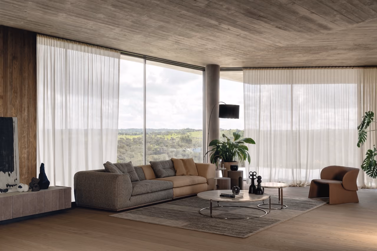 Modern living room with a large brown and gray sectional sofa, two round wooden coffee tables with decorative objects, a brown armchair, large green plants, and floor-to-ceiling windows with sheer white curtains overlooking a green landscape.
