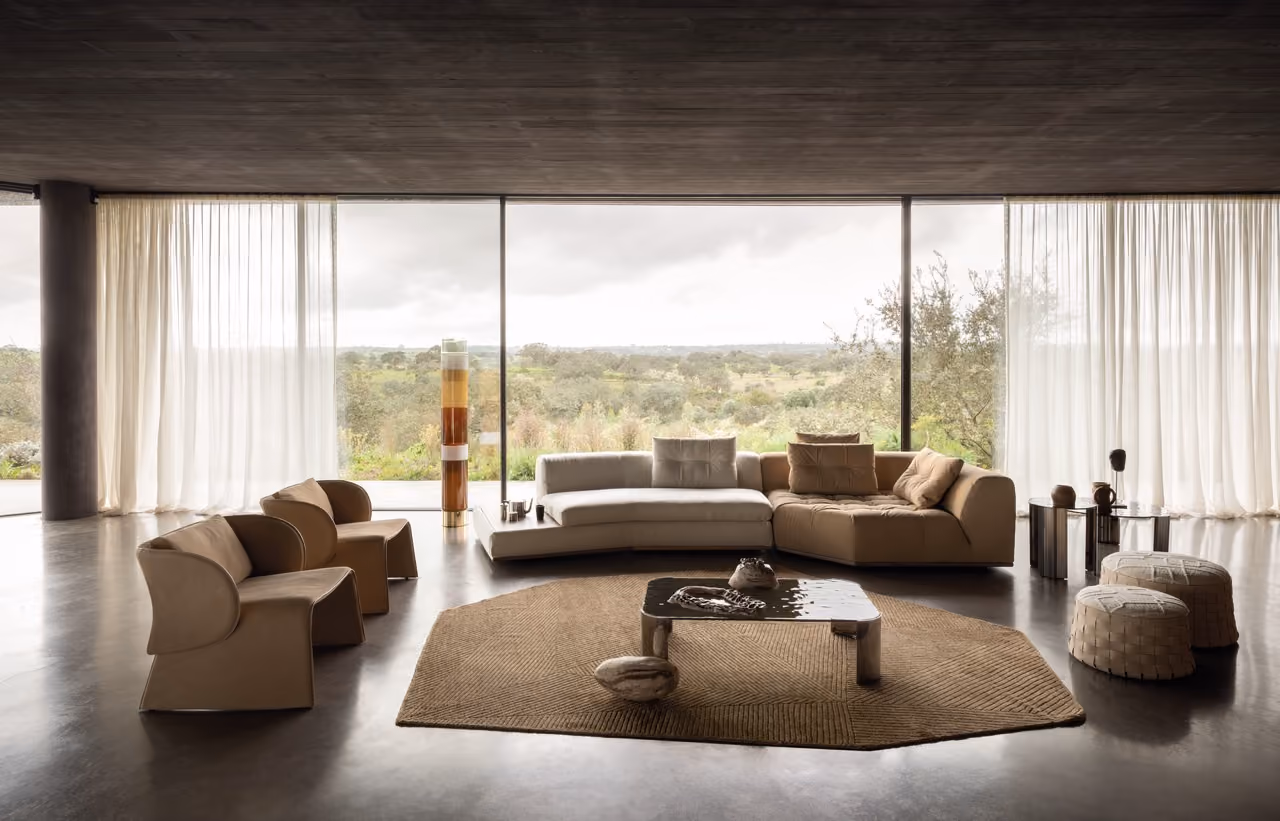 Modern living room with a large beige and brown sectional sofa, three brown armchairs, two woven poufs, a low rectangular coffee table on a textured brown rug, and floor-to-ceiling windows with sheer white curtains showing a green outdoor landscape.