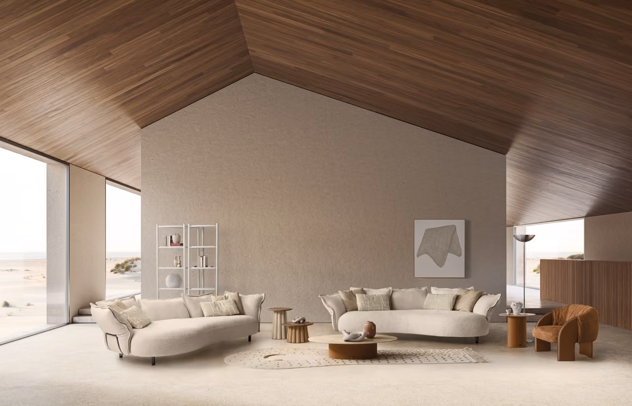Minimalist modern living room with two cream-colored curved sofas with beige cushions, small wooden side tables, an oval coffee table with decorative objects, a brown armchair, and a white shelving unit against a textured beige wall. The room has large windows showing a sandy outdoor landscape and a high angled wooden ceiling.