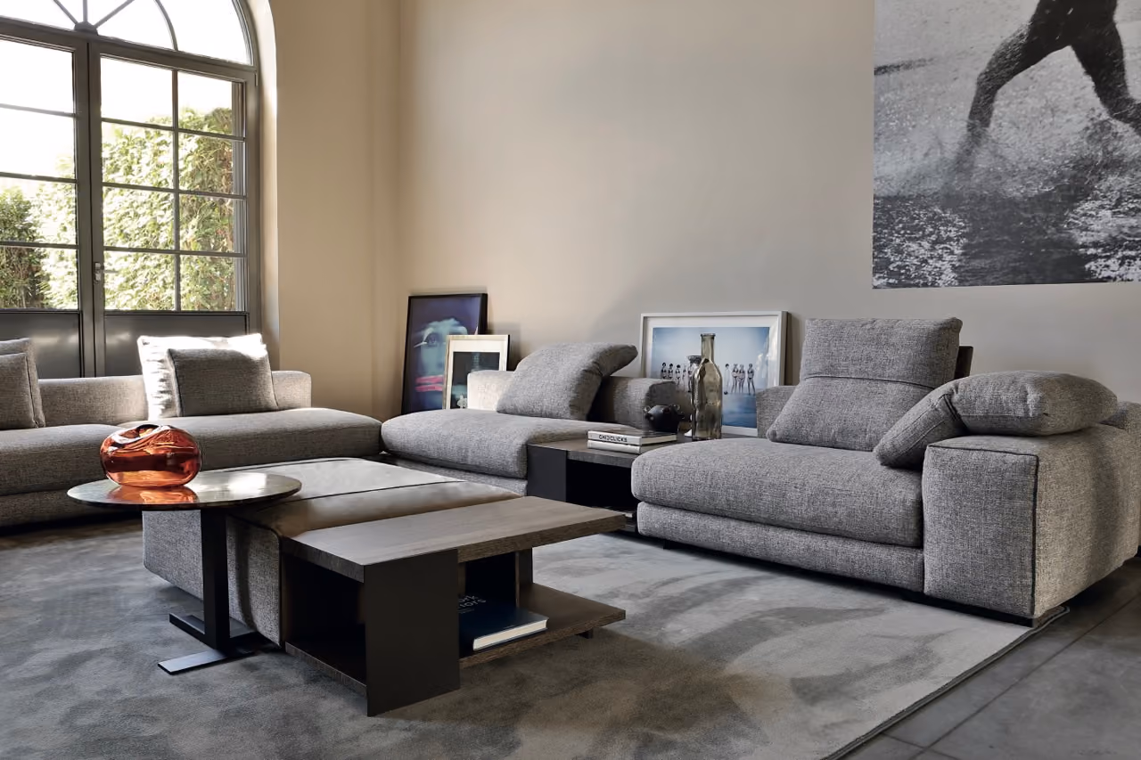 Modern living room with two gray fabric sofas and a matching armchair arranged around a modular dark wood coffee table and small round side table with a decorative glass bowl, set on a light gray area rug; large arched window lets in natural light, and framed art pieces lean against the beige wall.