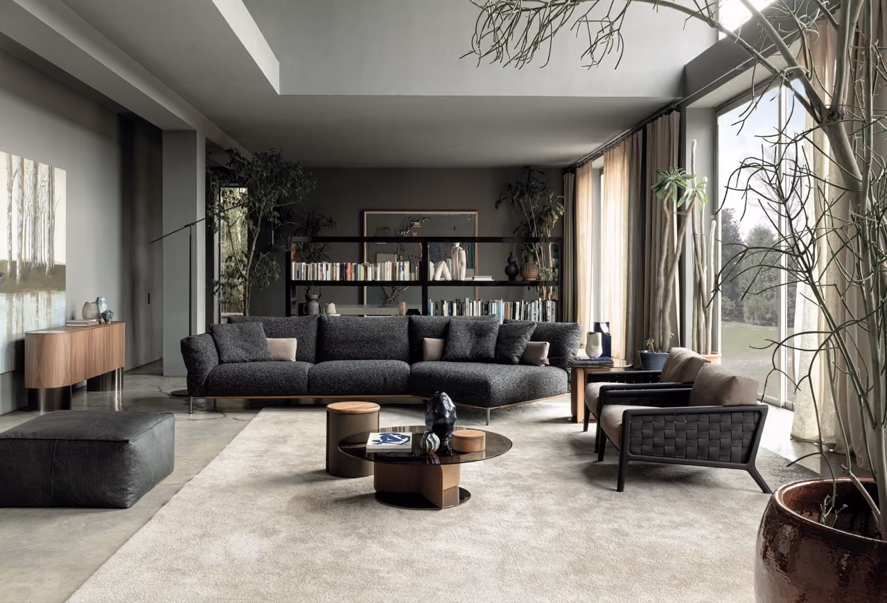 Modern living room with a large dark gray sectional sofa, two brown armchairs with black woven frames, and a black pouf on a light beige carpet. A round black coffee table with decorative objects is in front of the sofa. Floor-to-ceiling windows on the right bring in natural light, flanked by sheer curtains and tall green plants in pots. A wooden sideboard with decorative items and a large painting hang on the left wall, with a black bookshelf filled with books and vases behind the sofa.