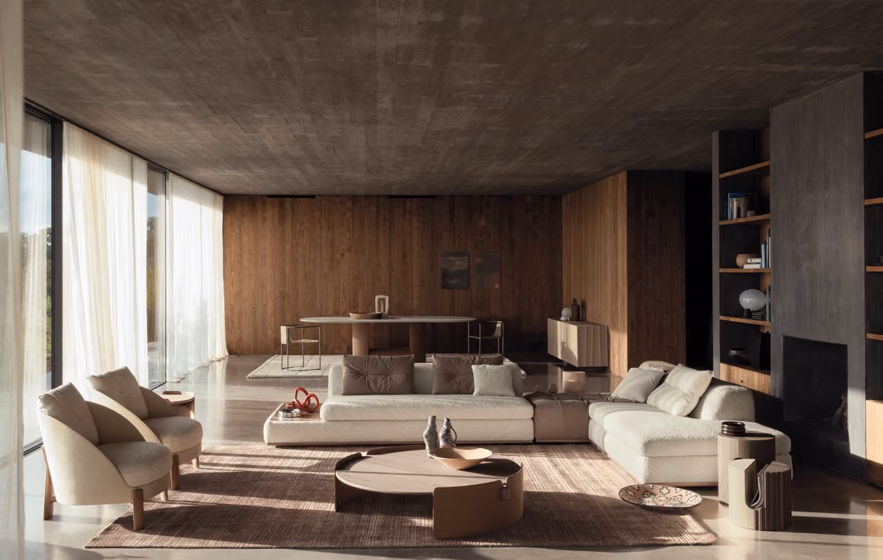 Modern living room with large floor-to-ceiling windows covered by sheer curtains, an L-shaped beige sofa with pillows, two rounded armchairs, a round wooden coffee table with decorative items, and a brown rug on a polished concrete floor; the background shows a wooden panel wall with a dining table and chairs, shelves with books, and a dark fireplace.