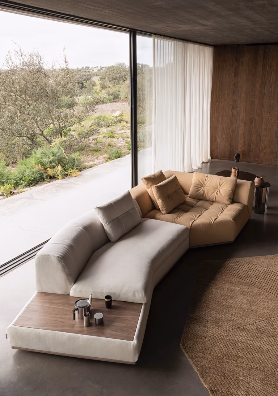Modern living room with an L-shaped sectional sofa featuring beige and light gray cushions, a built-in wooden side table with metal containers, large glass sliding doors overlooking a green landscape, sheer white curtains, a wooden accent wall, and a textured brown rug on a concrete floor.