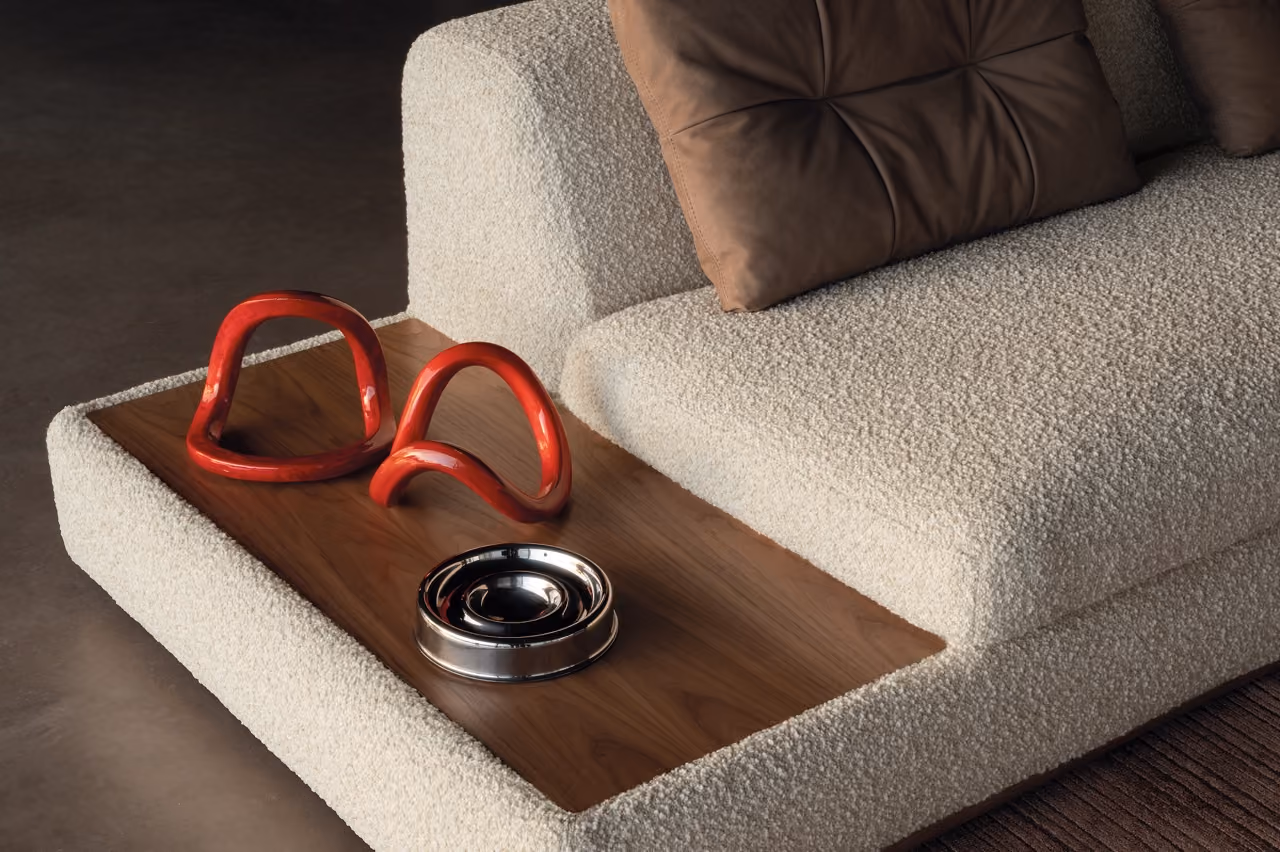 A close-up of a beige boucle sofa with brown cushions and an integrated wooden side surface holding two red ceramic decorative pieces and a round silver metal dish.