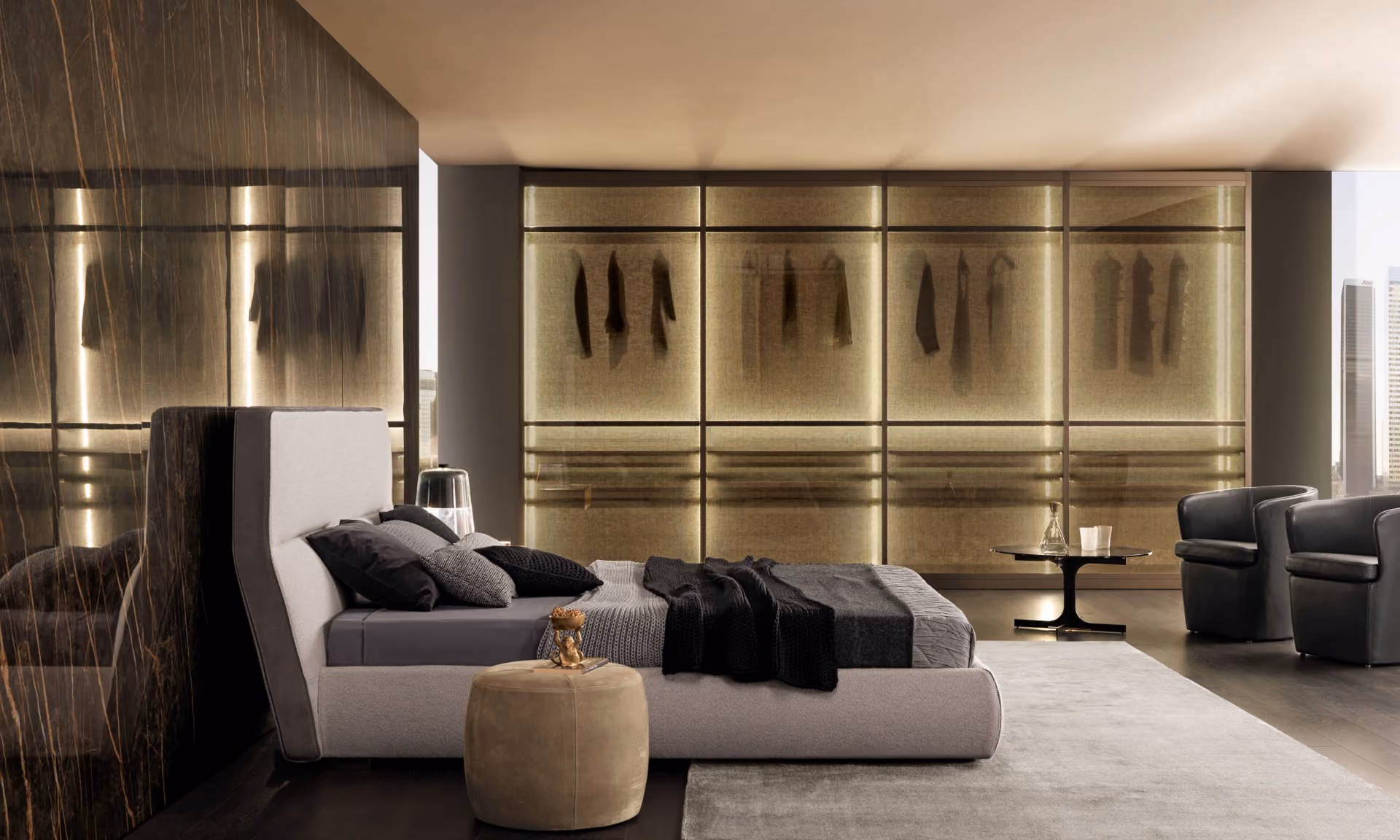 Modern bedroom with a low beige bed featuring gray and black bedding and pillows, a small round beige ottoman beside the bed, and a floor lamp. In the background, a large backlit closet with translucent panels displays hanging clothes silhouettes. On the right are two black armchairs and a small black round table with a glass decanter and two glasses. The room has dark wood flooring and a light gray rug.
