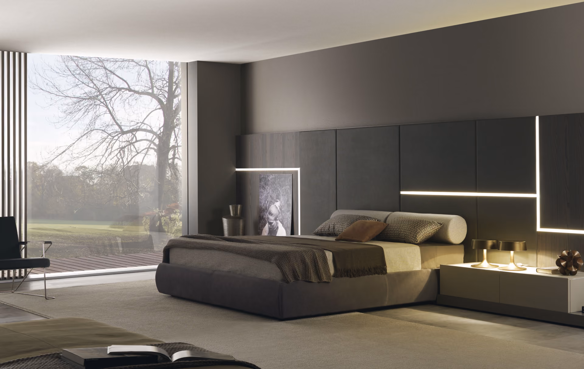 Modern bedroom with a large bed featuring a padded headboard with integrated geometric LED lighting, neutral colored bedding, two bedside lamps, a low white nightstand, a black framed photo leaning against the wall, a vase, and a large window showing a green outdoor scene.