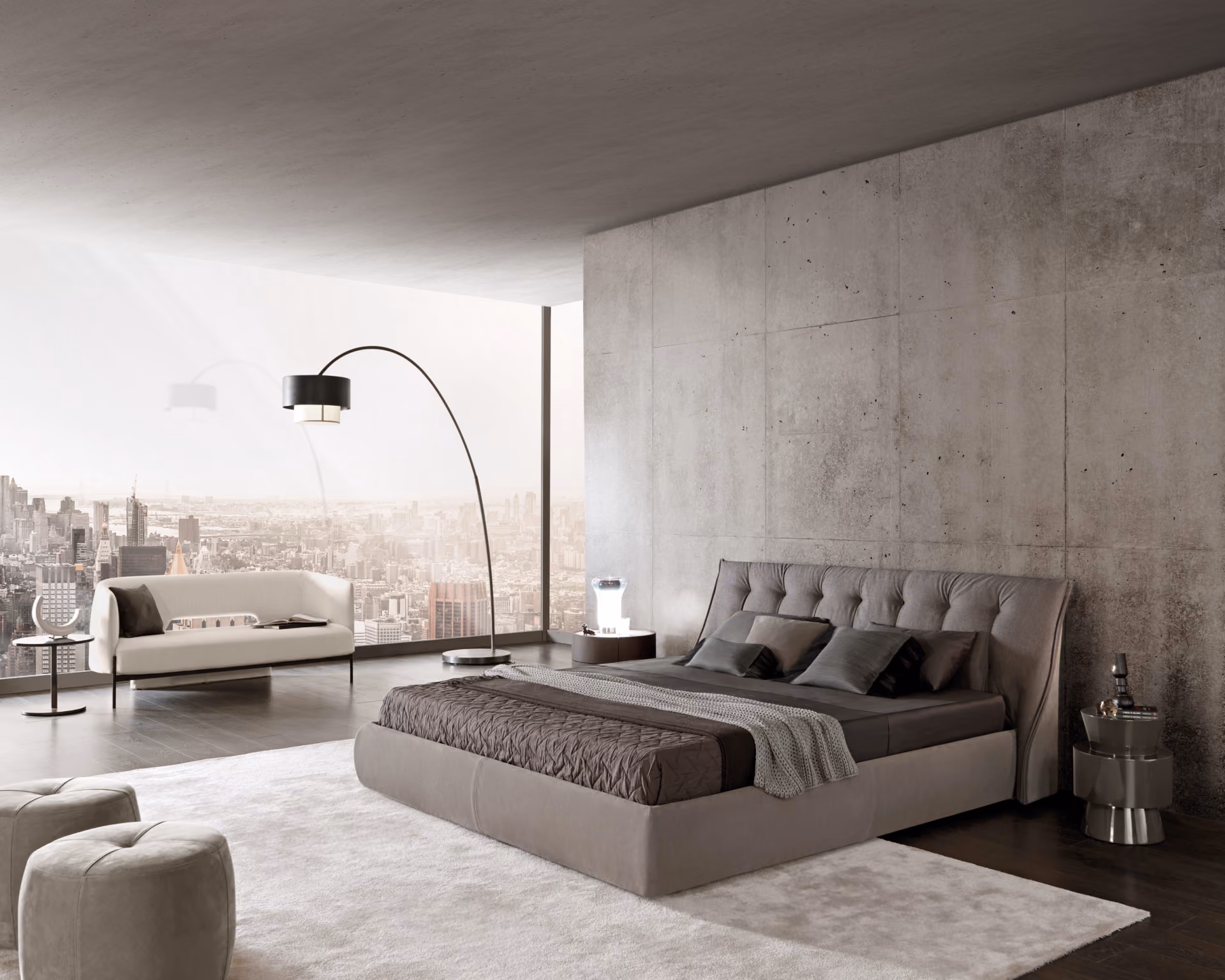 Modern bedroom with a large upholstered bed with multiple gray pillows and a brown quilt, a light beige sofa near a floor-to-ceiling window showing a city skyline, a tall curved floor lamp, a round side table with a modern lamp, and two beige ottomans on a white rug over dark wood floors.