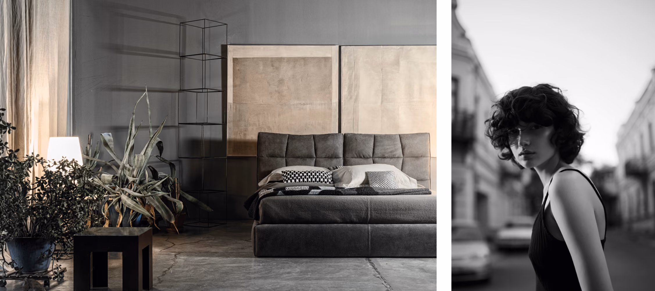 A modern bedroom with a gray upholstered bed featuring neutral pillows and a blanket, two large minimalist framed artworks on the gray wall behind it, a black metal shelving unit, potted plants, a glowing lamp, and a black side table on a concrete floor.