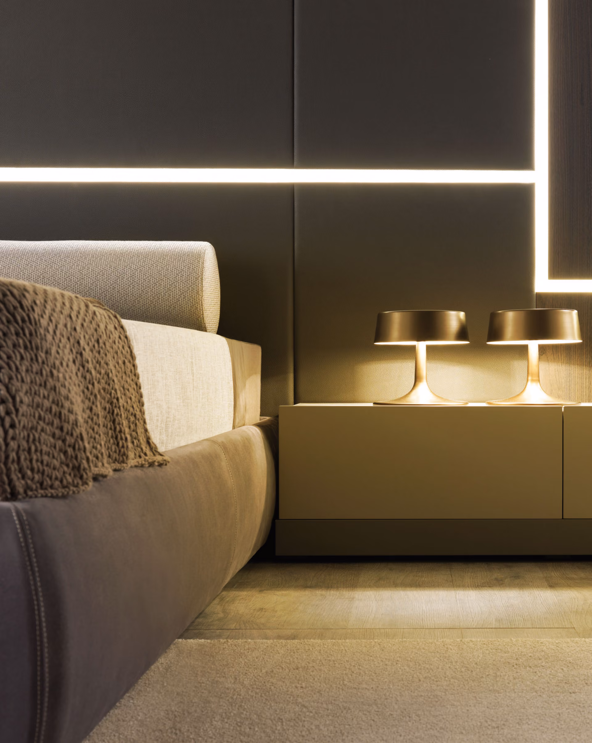Close-up of a modern bedroom corner showing a brown upholstered bed frame with beige bedding, a brown knitted blanket draped over the side, a beige nightstand with two contemporary bronze table lamps emitting warm light, and a wall with geometric light strips behind, complemented by wood flooring and a beige rug.