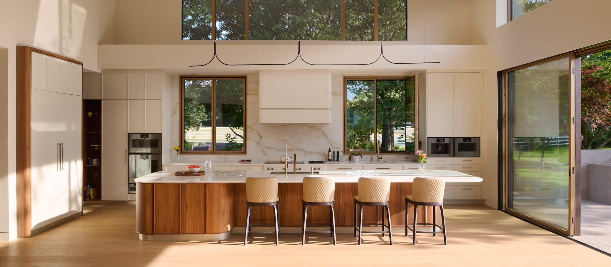 Modern spacious kitchen with a large wooden island featuring a white countertop, four beige cushioned chairs, built-in ovens on both sides, large windows showing greenery outdoors, and an open glass door leading to an outdoor area.