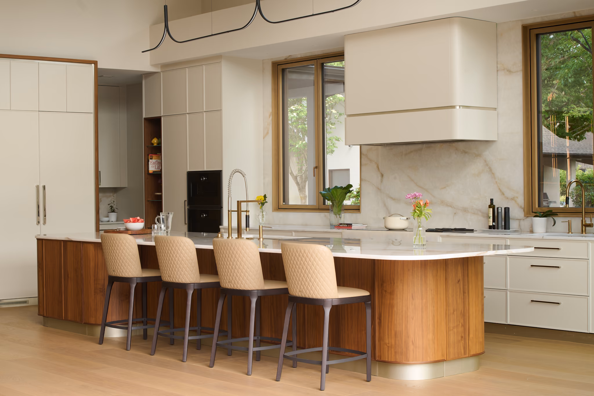 Modern kitchen with a wooden island topped with white marble, four upholstered beige bar stools, gold faucet, white cabinetry, large windows showing greenery outside, and a sleek white range hood above the stove.