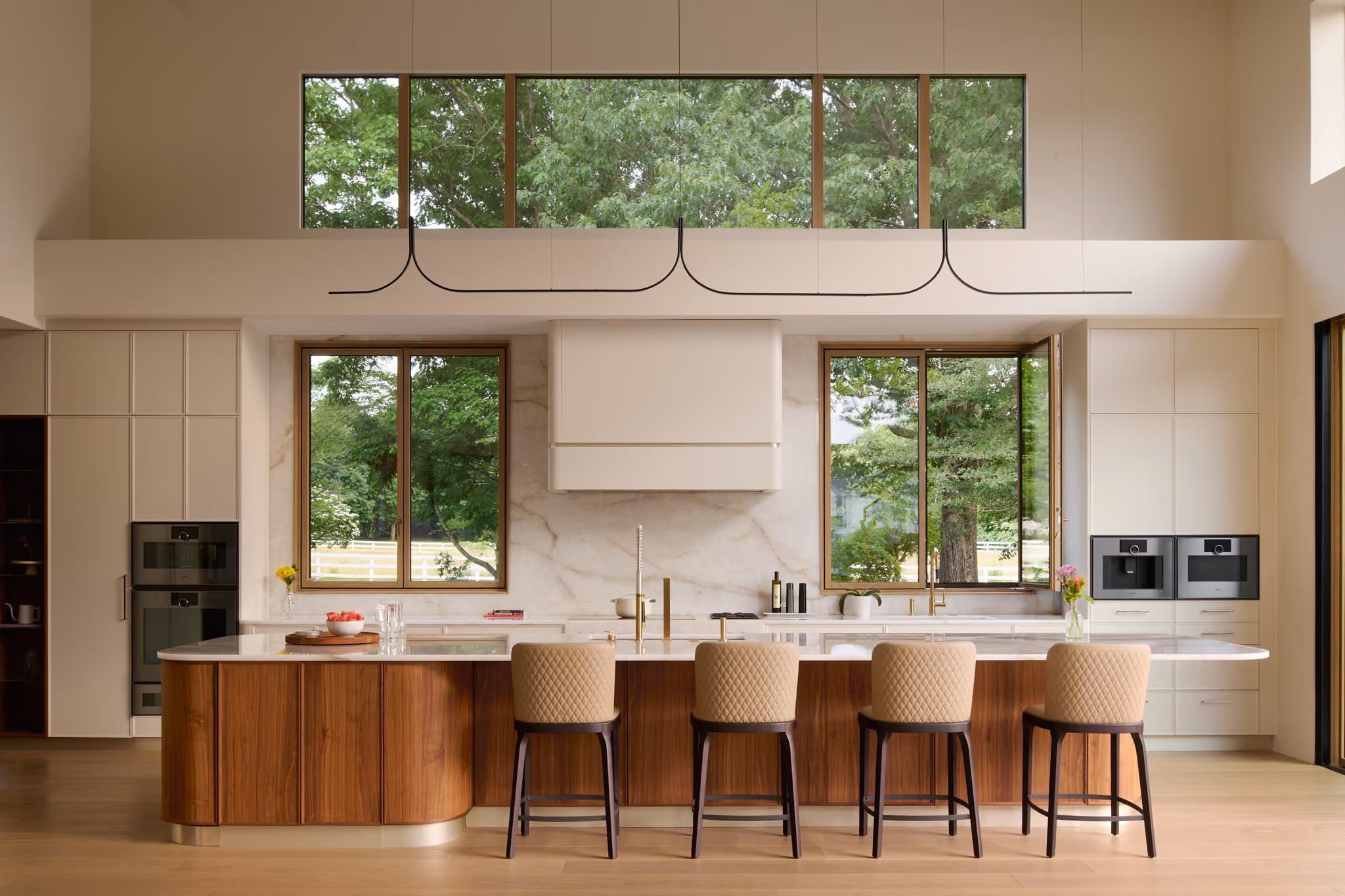 Modern kitchen with a large island featuring wood paneling and white countertop, four beige quilted bar stools, two windows showing green trees outside, white cabinetry with built-in black kitchen appliances, and a minimalist black pendant light above.