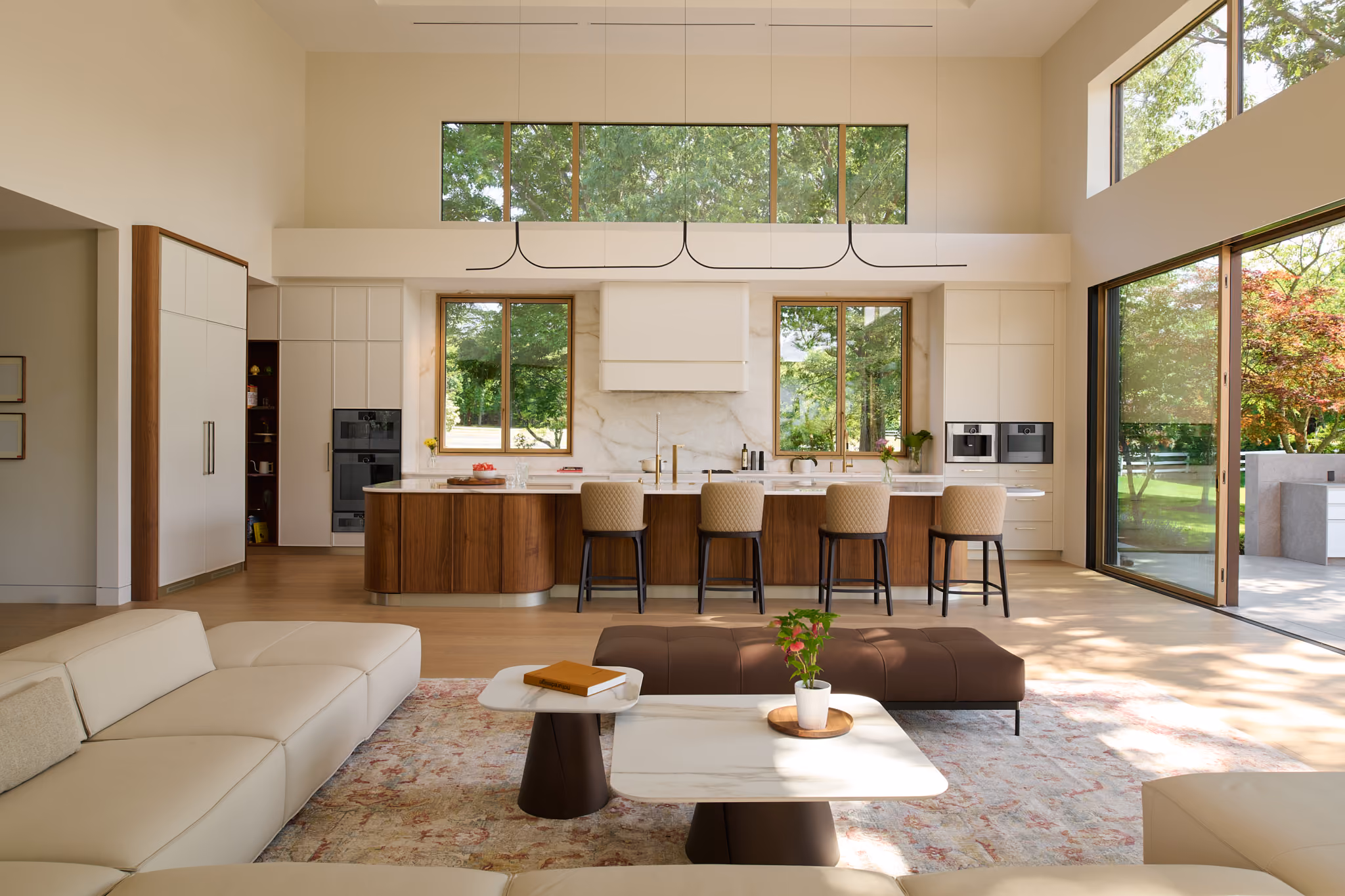 Modern open-concept kitchen and living area with large windows showing green trees outside, a central wooden island with four beige stools, marble countertops and backsplash, beige sectional sofa, brown ottoman, white marble coffee tables with plants and books, light wood flooring, and sliding glass doors leading to a patio.