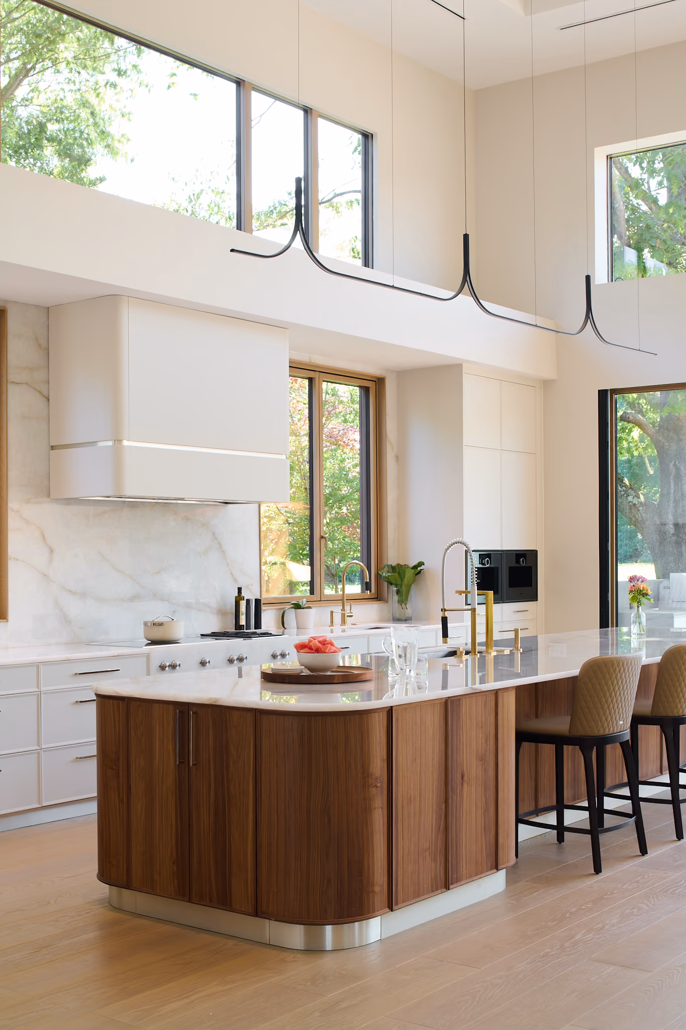 Modern kitchen with large wooden and white marble island featuring two high chairs, gold-tone faucets, a bowl of cut watermelon, and large windows letting in natural light and showing green trees outside.