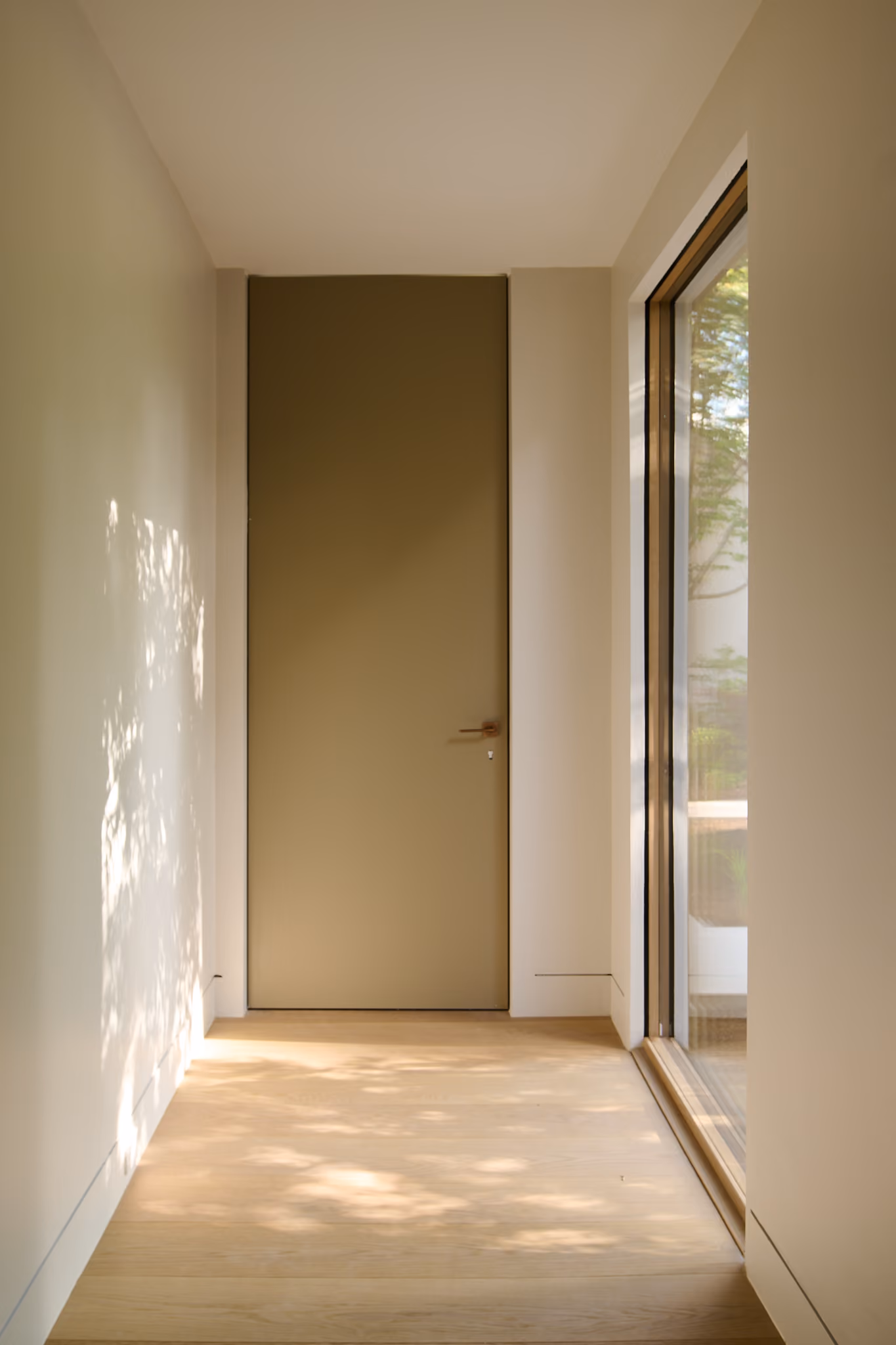 Minimalist hallway with light wooden floor, a closed simple khaki-colored door at the end, and a large window on the right letting in sunlight that casts tree shadows on the floor and left wall.