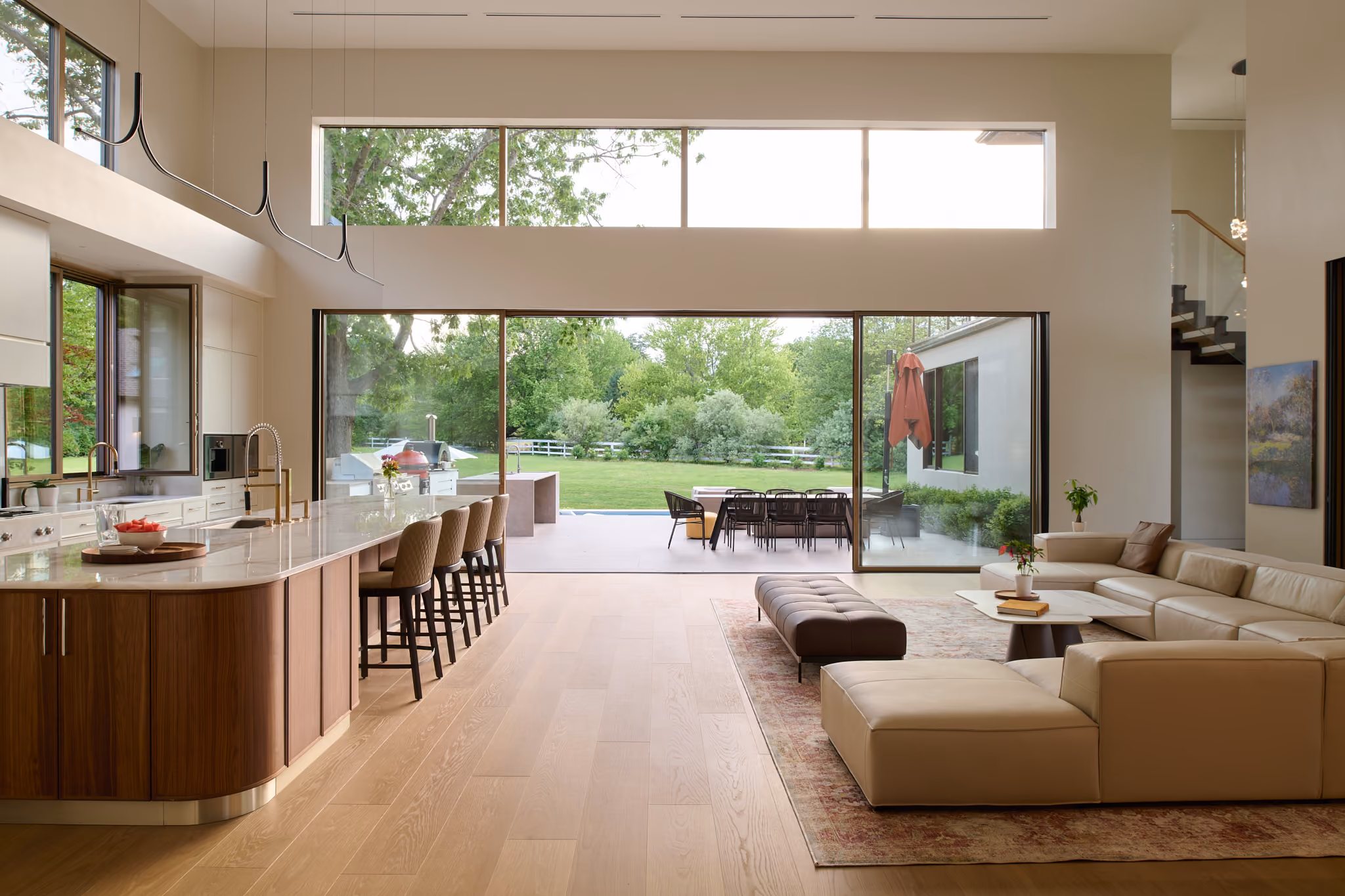 Modern open-plan living space with a large kitchen island featuring a white marble countertop and five beige cushioned stools, a beige sectional sofa on a patterned rug, a brown leather ottoman, and a sliding glass door opening to a patio with a dining table and chairs, overlooking a green yard with trees and a white fence.
