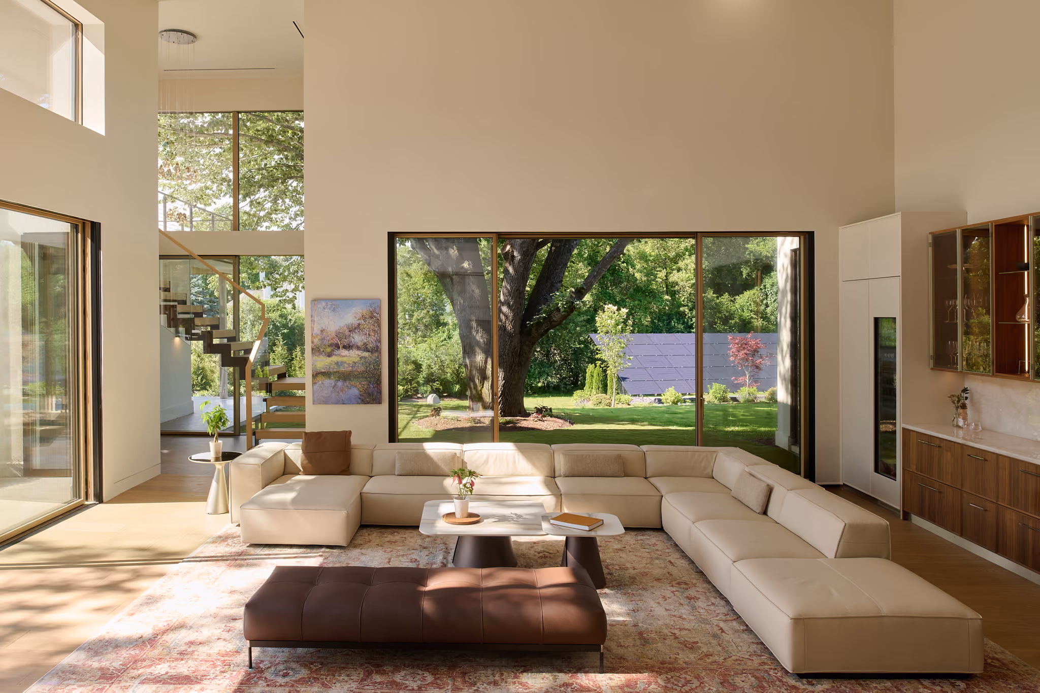 Spacious modern living room with a large beige sectional sofa, two white coffee tables with plants and books, a brown leather ottoman on a patterned rug, and floor-to-ceiling windows showing a green garden with trees and solar panels.