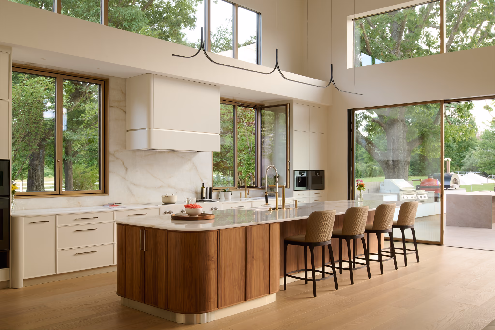 Modern kitchen with a large marble countertop island featuring wooden base, four upholstered bar stools, and gold faucets, surrounded by white cabinetry and large windows showing green trees outside.