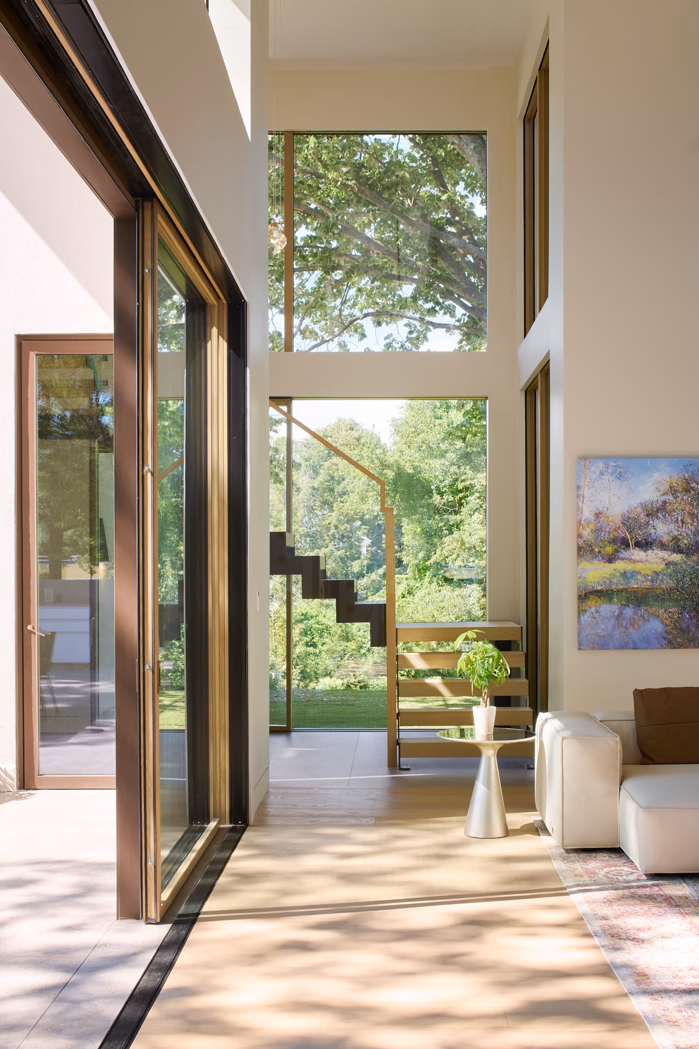 Bright modern living room with large floor-to-ceiling windows showing lush green trees outside, a wooden staircase with glass railing, a small round side table with a potted plant, and a white couch partially visible on a patterned rug.