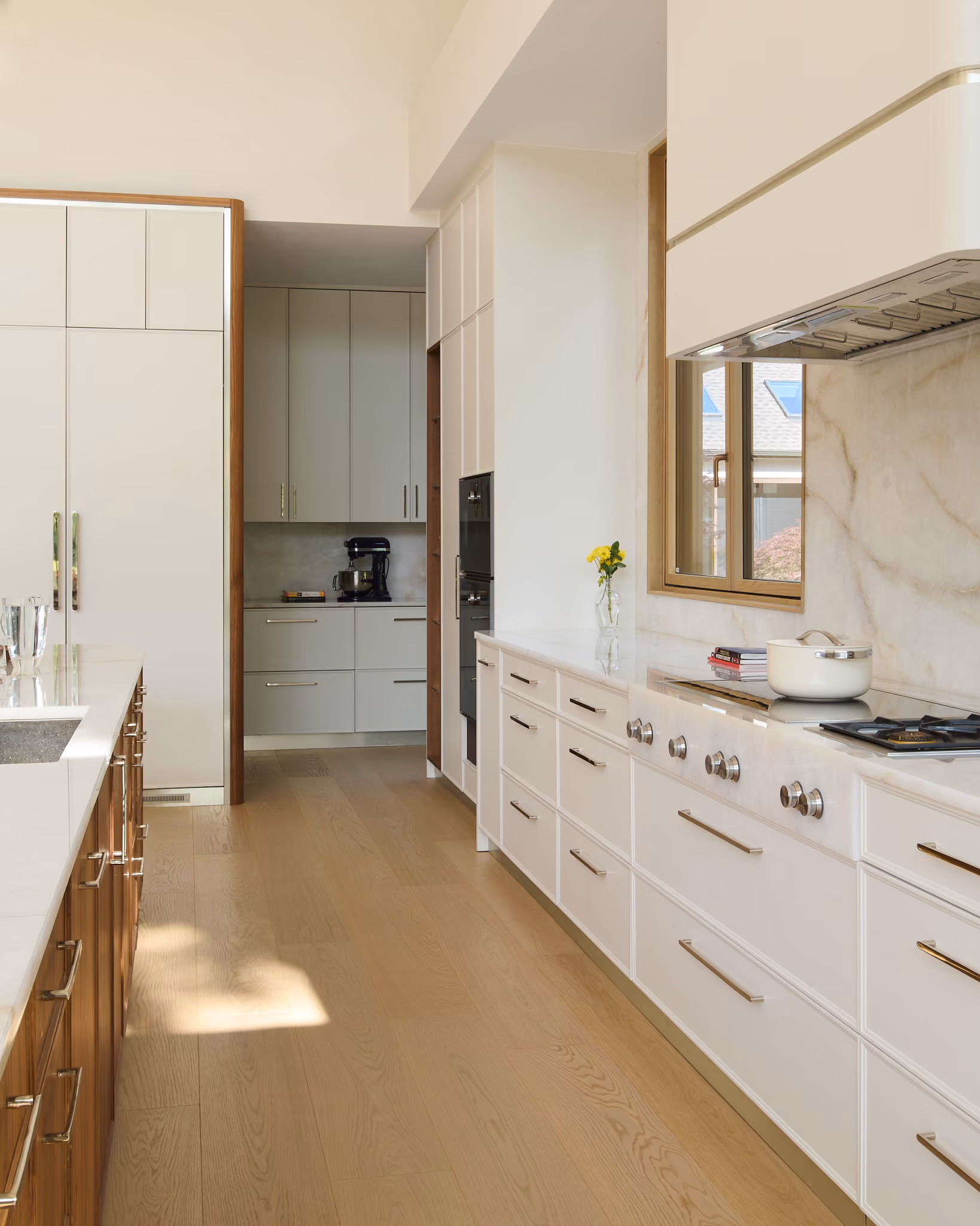 Modern kitchen with light wood flooring, white cabinets, and marble countertops; features a stovetop with a white pot, a window overlooking houses, an island with a sink on the left, and a small vase with yellow flowers near the window.