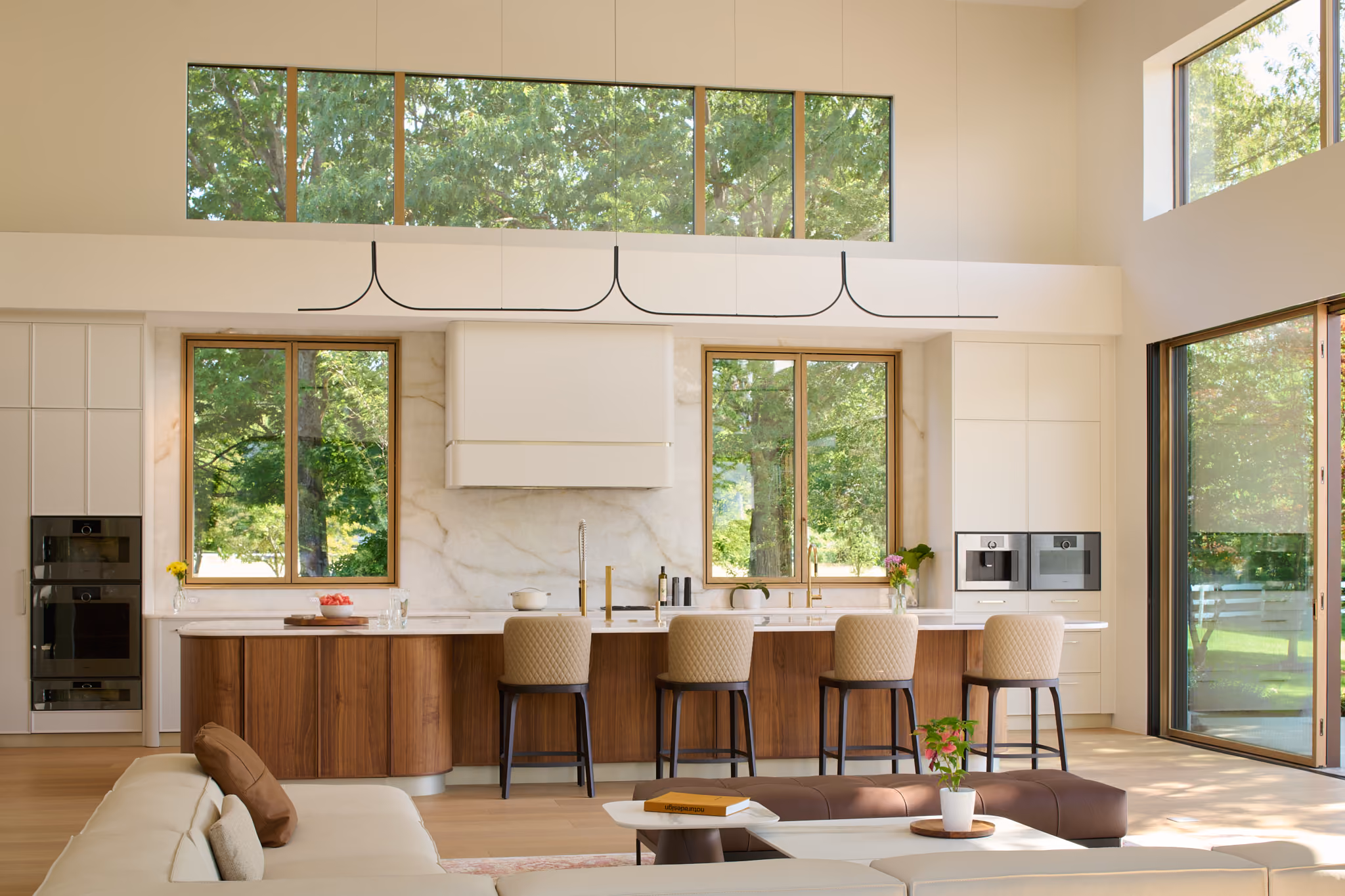 Modern kitchen with large windows showing green trees outside, a long marble-topped island with wooden cabinets, four beige quilted bar stools, built-in ovens and coffee machine, and an adjacent living area with a beige sofa, coffee table with a book, and a plant.
