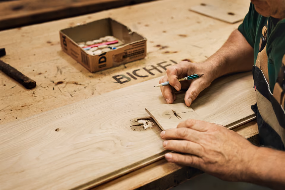 A person working on a wooden plank with visible knots, using a pencil and a small wooden piece to mark or measure on a workbench with a box of chalk or markers nearby.