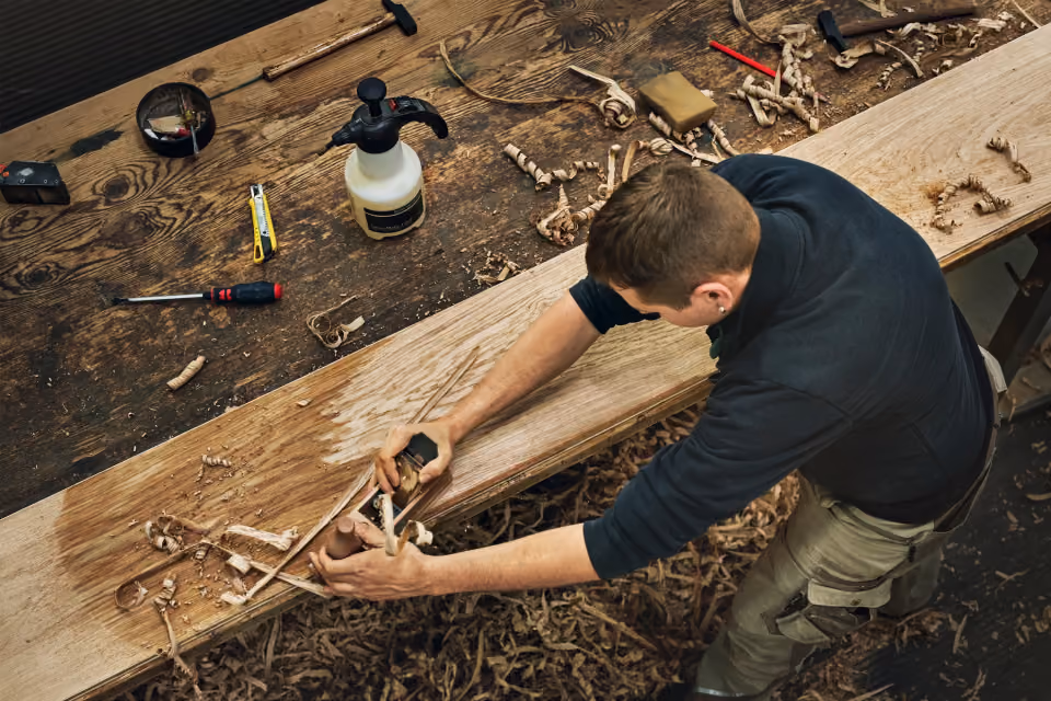 A man using a hand plane tool to smooth the surface of a wooden plank on a workbench with wood shavings scattered around.