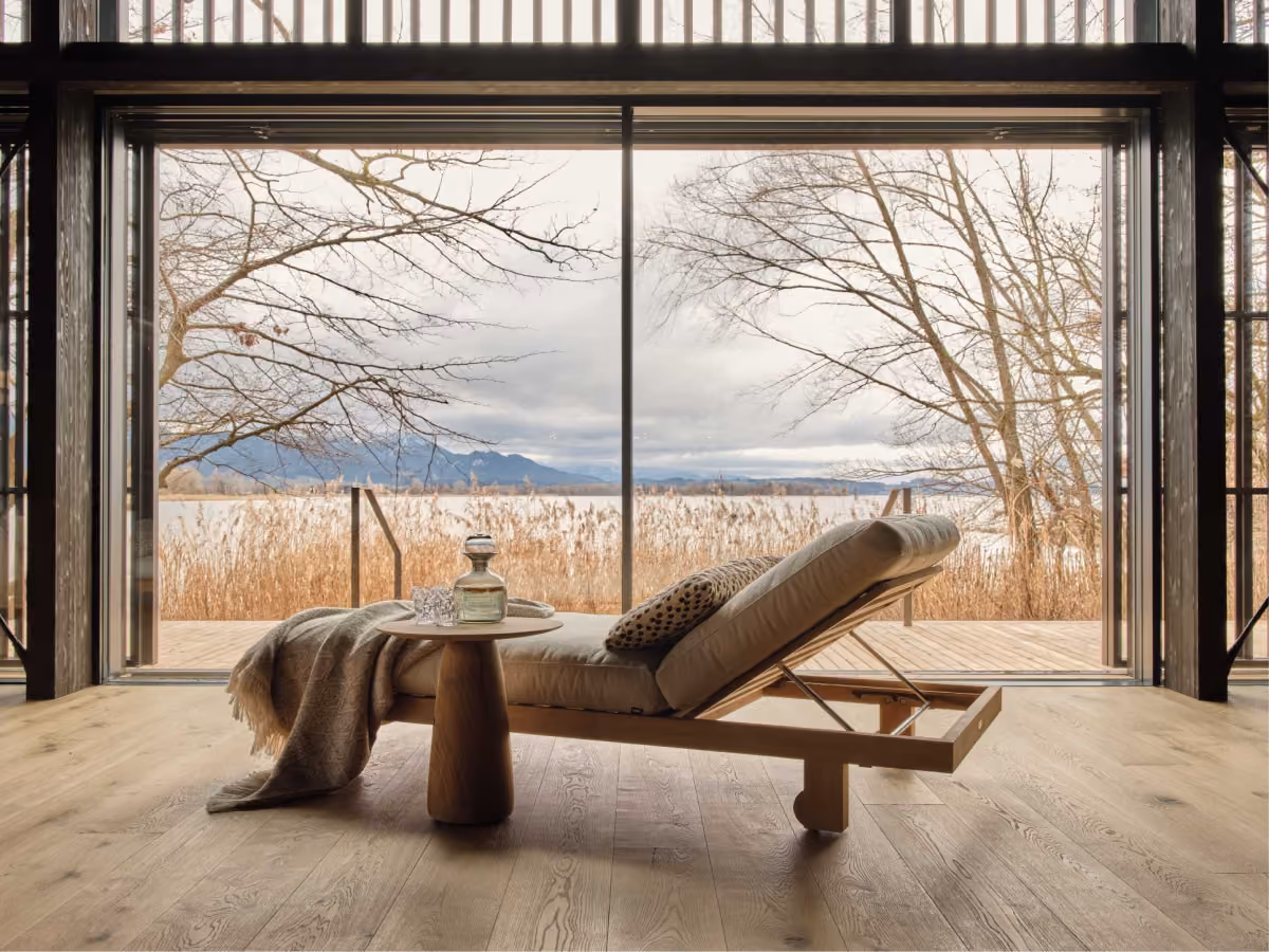 Minimalist lounge chair with beige cushions and a throw blanket beside a small wooden side table holding a glass decanter and two glasses, set inside a room with large sliding glass doors overlooking a lakeside view with tall reeds, leafless trees, and distant mountains under a cloudy sky.