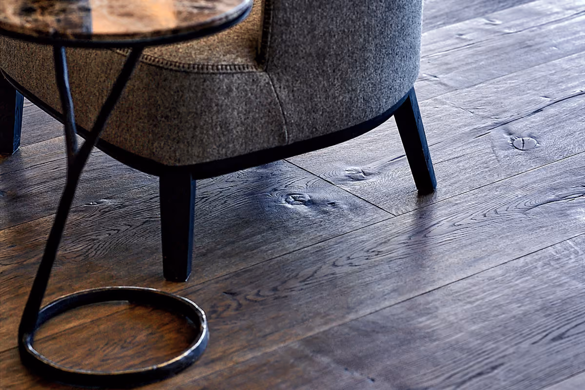 Close-up of a textured gray upholstered chair with black legs on a wooden floor with visible grain and knots, beside a small round side table with a slender black metal support.