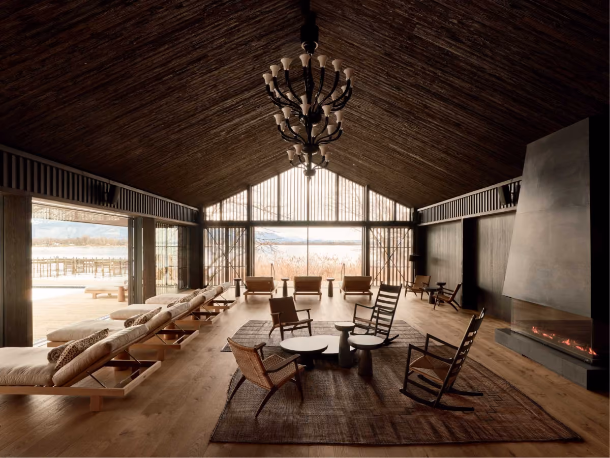 Modern lounge interior with dark wood vaulted ceiling, large windows overlooking lake and mountains, a row of cushioned lounge chairs on the left, wicker and wooden armchairs around a central coffee table on a rug, and a lit fireplace on the right.