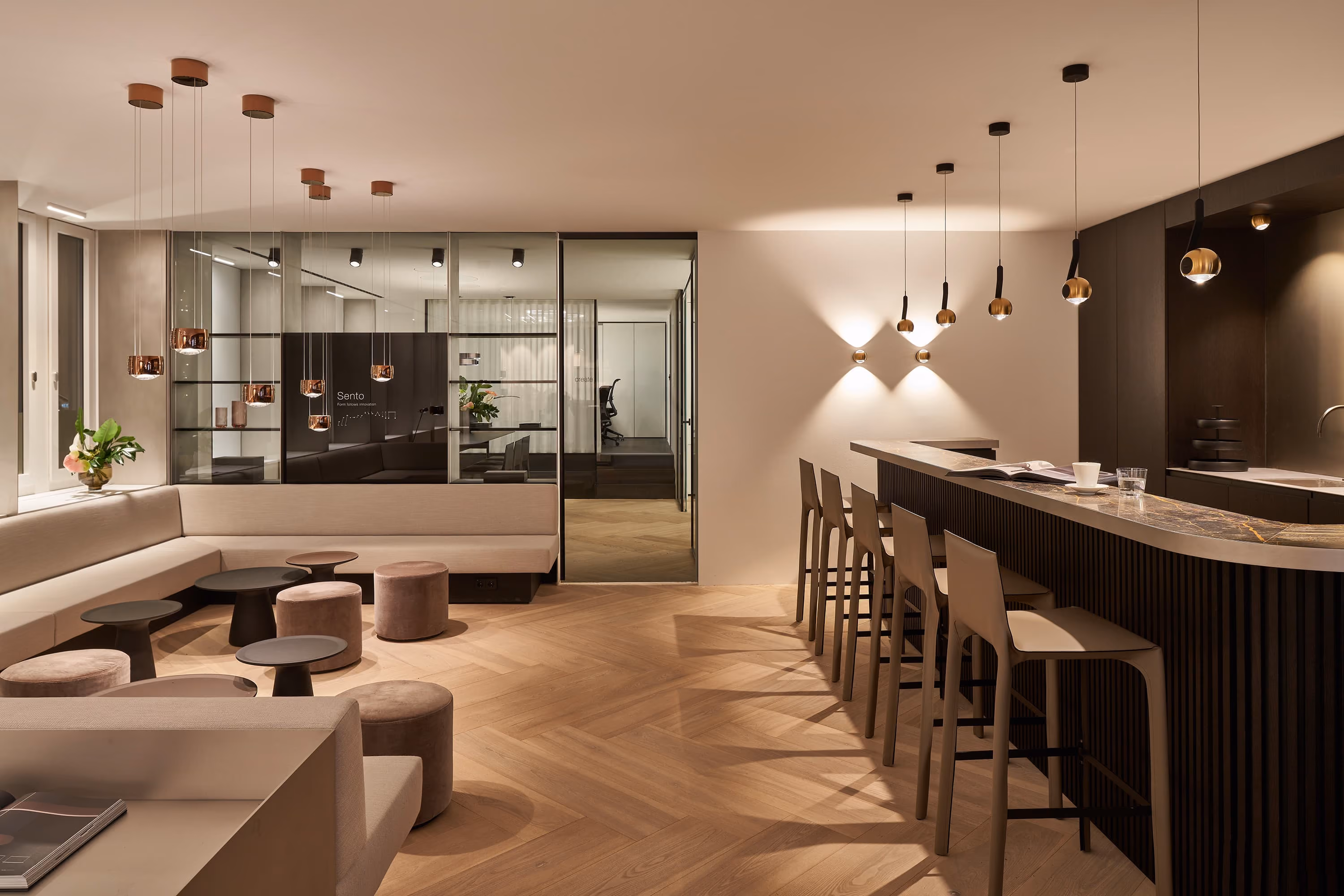 Modern, minimalist lounge area with beige sectional sofa, round stools, small black tables, brass pendant and wall lights, and a dark wood bar counter with five beige bar stools on light wood flooring.