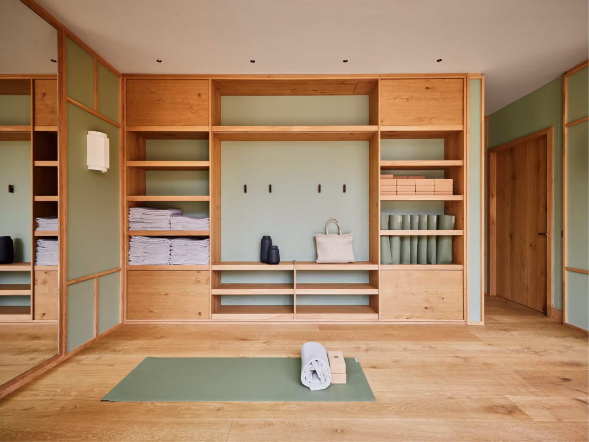 Minimalist yoga room with wooden floor, green yoga mat with a rolled towel and two wooden blocks on it, and built-in wooden shelves holding folded towels, yoga blocks, rolled mats, and decorative vases and a woven bag on the bench.