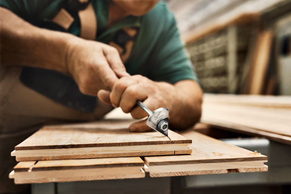 A woodworker carving or smoothing a layered wooden plank using a metal hand tool in a workshop.