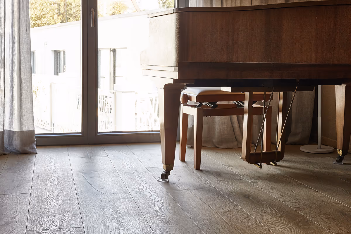 Lower section of a wooden grand piano with brass pedals on caster wheels, positioned near a large glass door with curtains and natural light reflecting on the wooden floor.