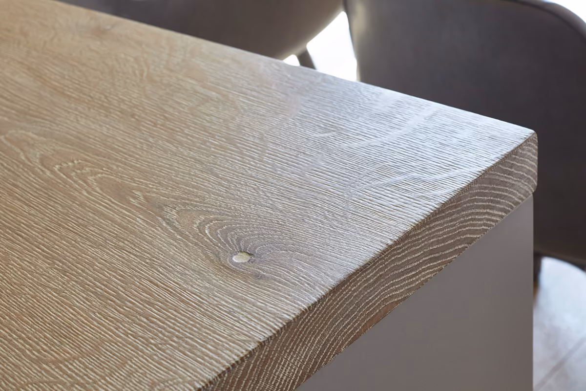 Close-up of a light wooden table corner with visible wood grain texture and a smooth finish, with blurred dark chairs in the background.