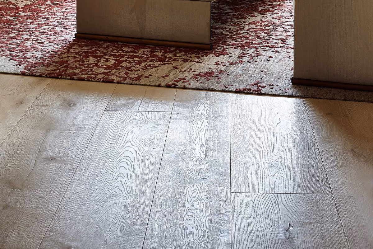 Close-up of light wooden floorboards with visible wood grain texture, partially covered by a red and beige patterned rug near the base of a door frame.