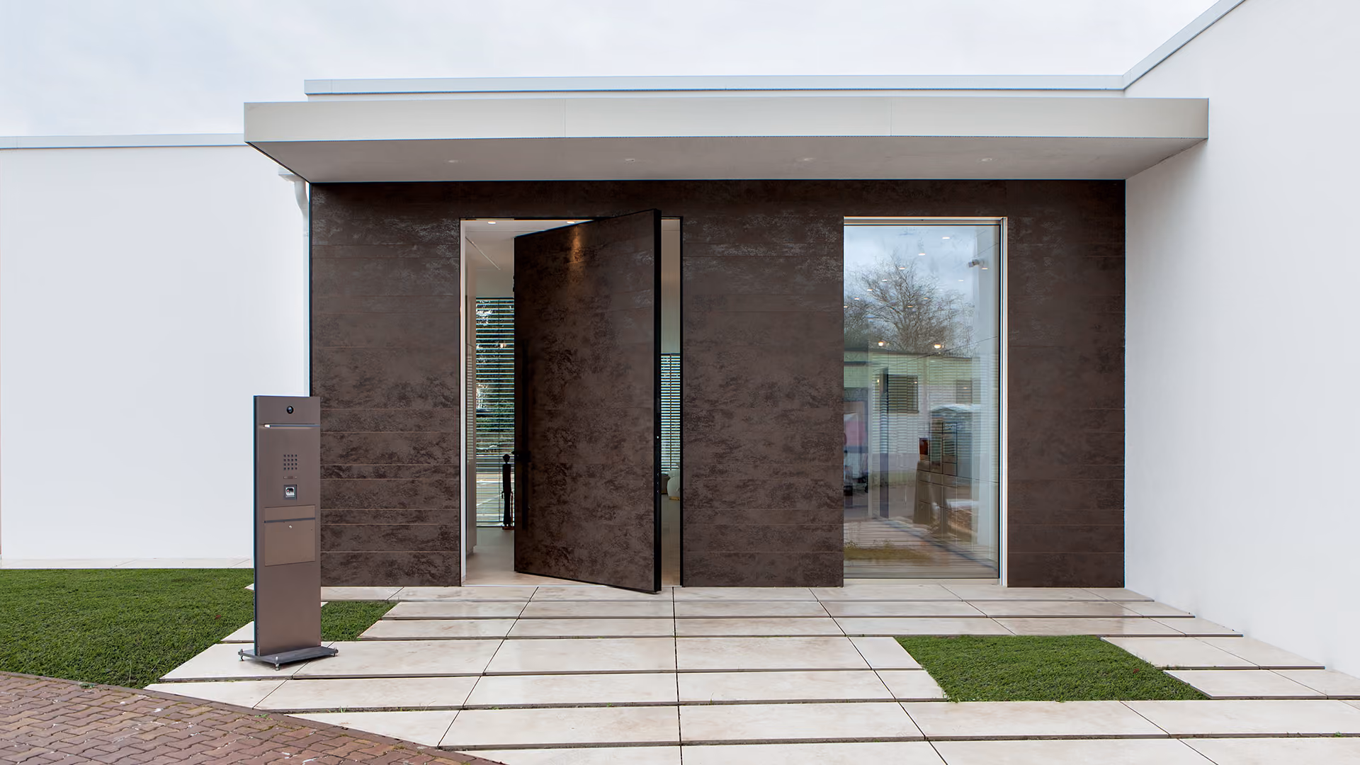 Modern building entrance featuring a large, dark pivot door partially open, flanked by a tall window and white walls, with tiled flooring and patches of green grass.