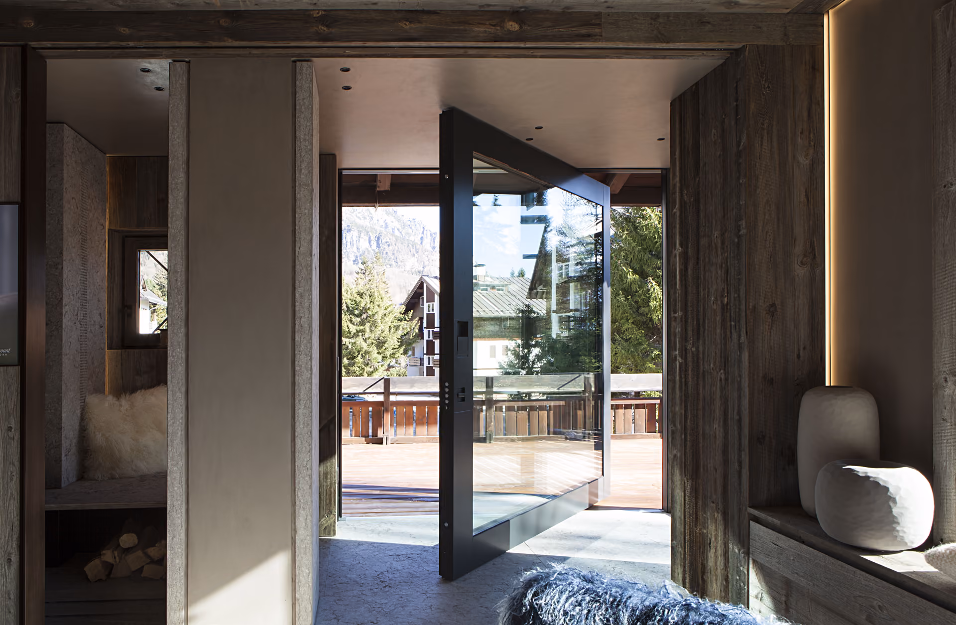 Interior view of a modern rustic home featuring a large glass pivot door opening to a wooden deck with outdoor scenery including trees and a mountain in the background.