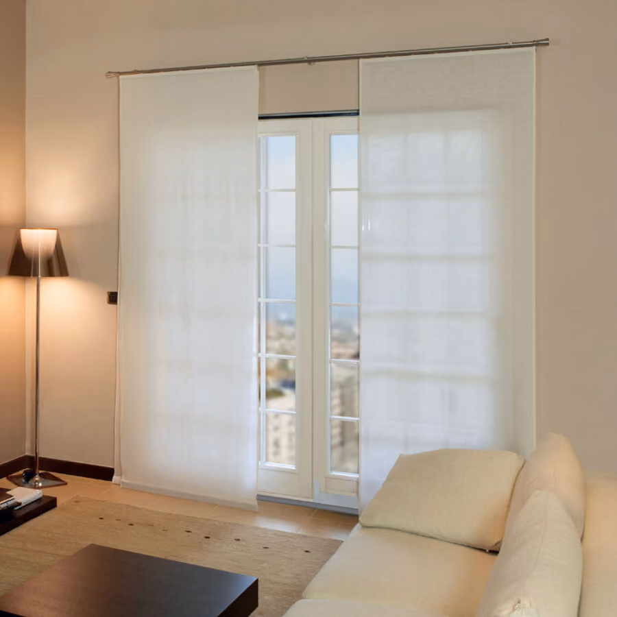 Modern living room with beige walls, a cream fabric sofa, dark wooden coffee table, tall floor lamp with a conical shade, and two light sheer sliding curtains covering glass doors overlooking a city view.