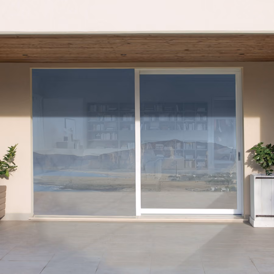 Modern sliding glass door with white frame reflecting an outdoor landscape and showing a blurred bookshelf inside, flanked by two potted plants on a tiled porch.