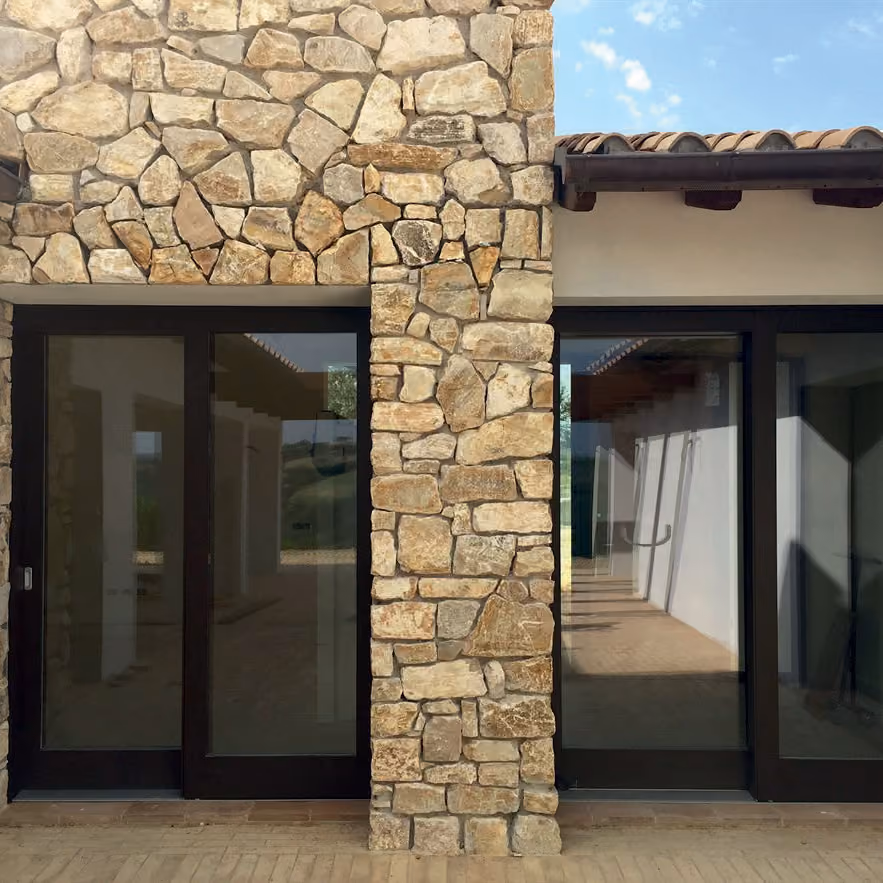 Two large sliding glass doors framed by black borders set in a stone wall with irregularly shaped beige and brown stones, with a tiled floor in front and part of a roof with exposed wooden beams above.